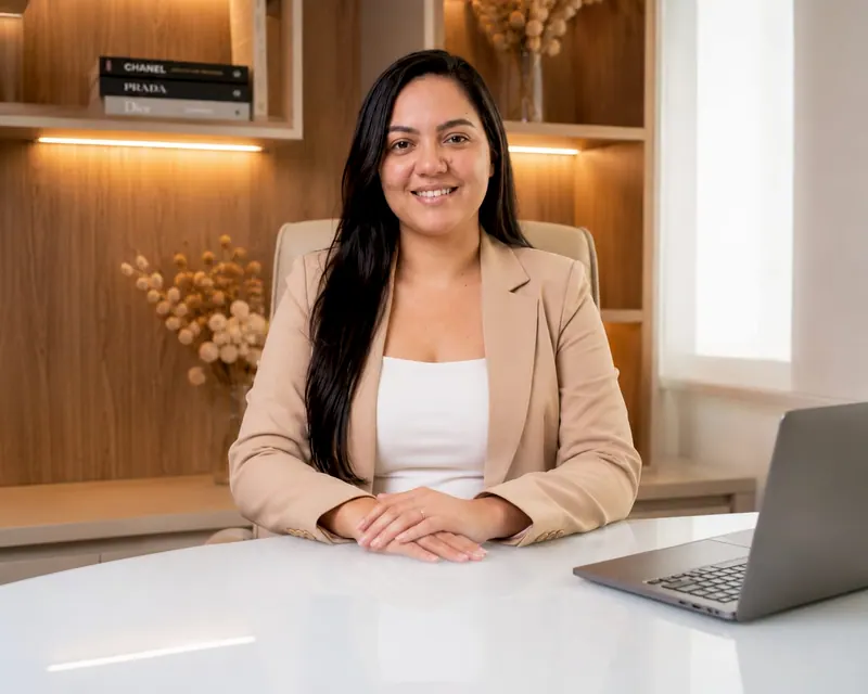 Dayane de blazer bege sentada à mesa branca do escritório, com notebook e estante com luz quente ao fundo