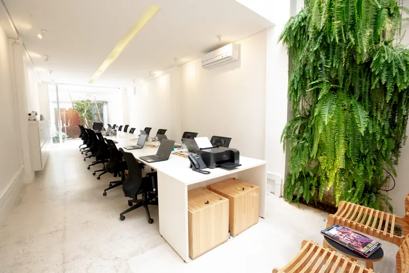 Sala ampla do escritório com mesas de trabalho, parede de plantas e luz natural
